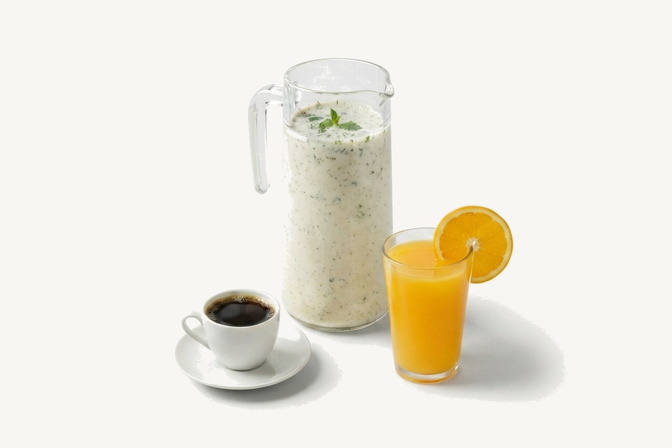 A glass jug of borhani flecked with mint, a glass of orange juice with an orange slice, and a white cup of black coffee on a saucer