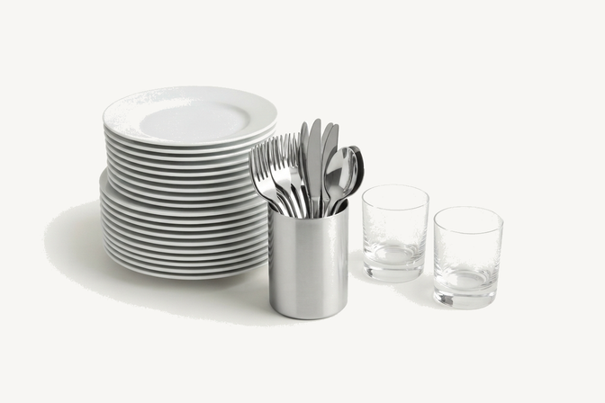 A stack of white plates, cutlery standing in a steel holder, and two water glasses