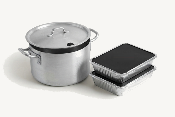 An aluminium dekchi with its lid on, beside two stacked foil trays with plain black lids