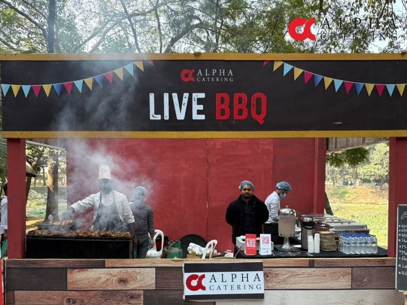 A cook grilling skewers over smoke at the Alpha Catering live BBQ stall, with crew serving at the counter beside him